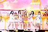 A girl group performs on a colorful pastel stage with a lemonade stand backdrop.