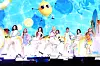 Hearts2Hearts members perform in colorful outfits on a bright blue stage with beach-themed graphics.